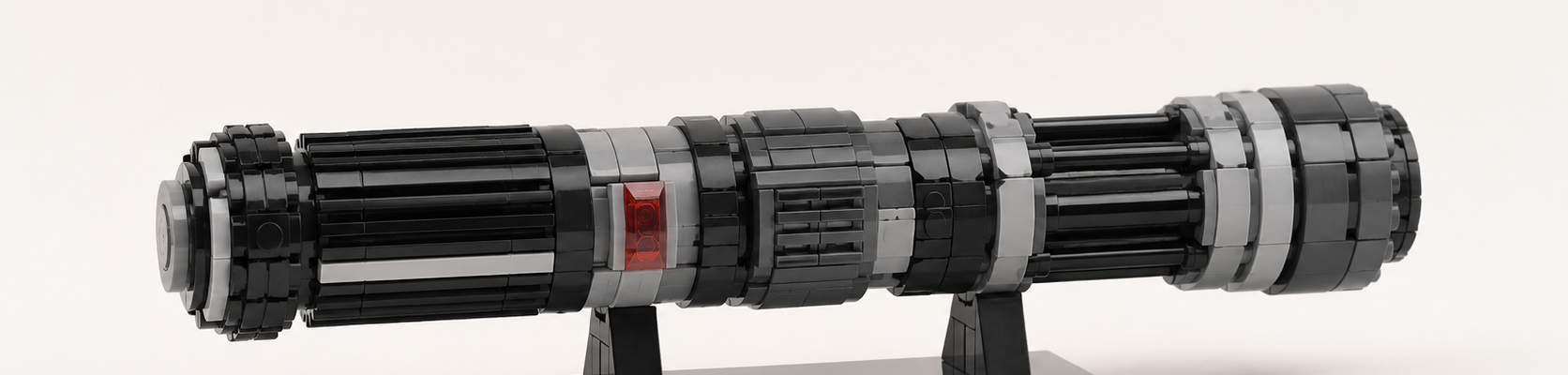 Generyczna klockowa rękojeść sci-fi w czerni i szarościach z małym czerwonym przezroczystym detalem, ustawiona na prostej podstawce na jasnym neutralnym tle.